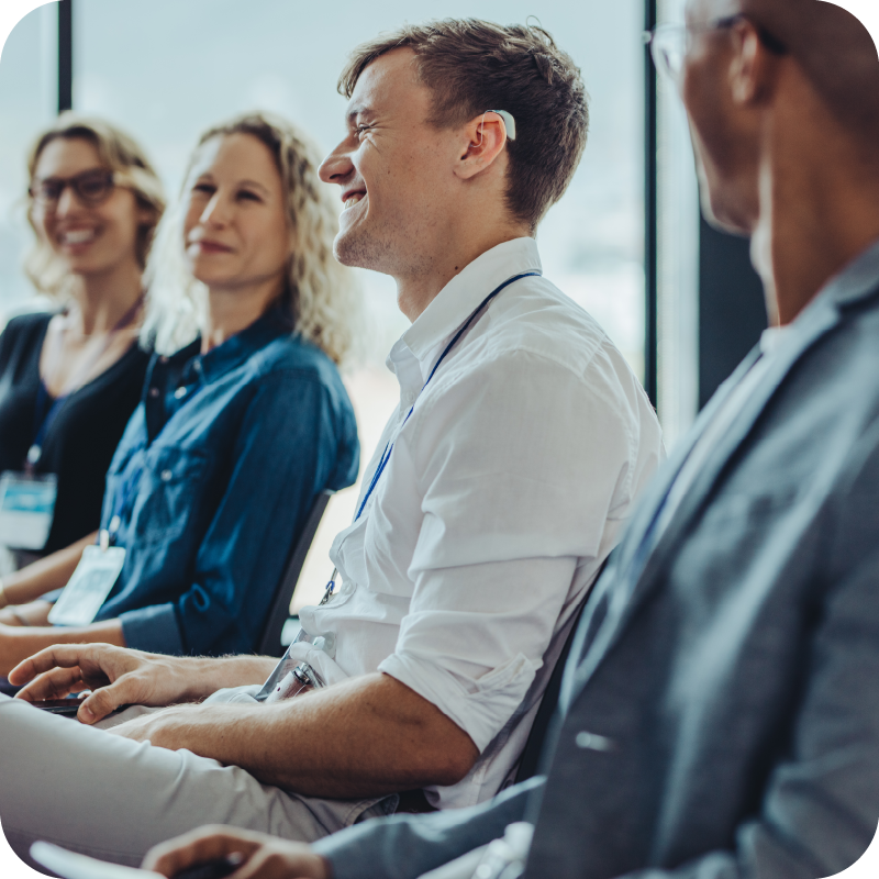 Professionals in business attire sit together in a meeting; a Bluetooth enabled hearing aid is worn by one attendee who is smiling and engaged with coworkers.